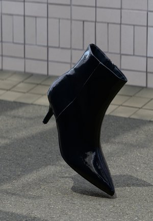 Bottine noire en cuir verni avec talon aiguille, posée sur une surface pavée devant un mur carrelé.