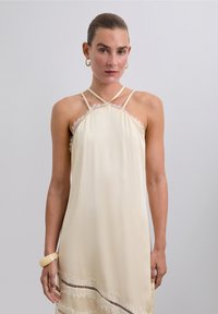 Robe de couleur crème en tissu lisse, dotée de franges délicates le long de l'encolure et de l'ourlet, avec des bretelles fines et de légères rayures horizontales en bas.