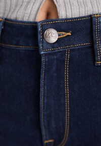 Jeans in denim blu scuro con cuciture dorate. Presentano un bottone metallico con il logo "Lee" e un design a vita alta. Il tessuto texturizzato è visibile.