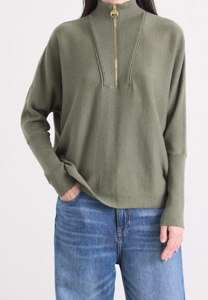 Femme portant un pull vert olive avec une fermeture éclair dorée à moitié et un jean bleu, une main dans la poche et l'autre détendue sur le côté.