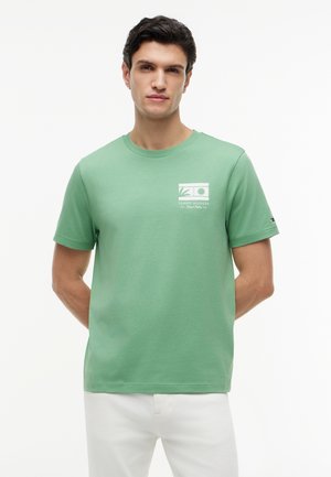 Ung mann står vendt fremover, iført en grønn Tommy Hilfiger t-skjorte med hvitt trykk på brystet og hvite bukser, hendene bak ryggen.