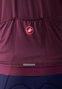 Haut de sport bordeaux en tissu texturé, avec un logo rond et une bande réfléchissante à l'ourlet. Ajustement extensible avec un dos froncé.