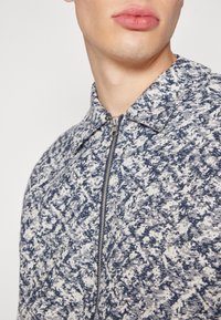 Strikket lynlåstrøje i marineblå og hvid med krave og tekstureret, meleret mønster. Nærbillede viser detalje af lynlåsen og stoftekstur.