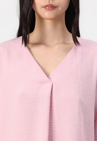 Lys rosa bluse med V-hals, med en teksturert stoff og en plissering ved halslinjen.