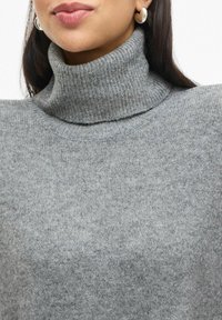 Maglione grigio a collo alto con texture a costine, realizzato in un morbido materiale a maglia e con lievi variazioni di colore.