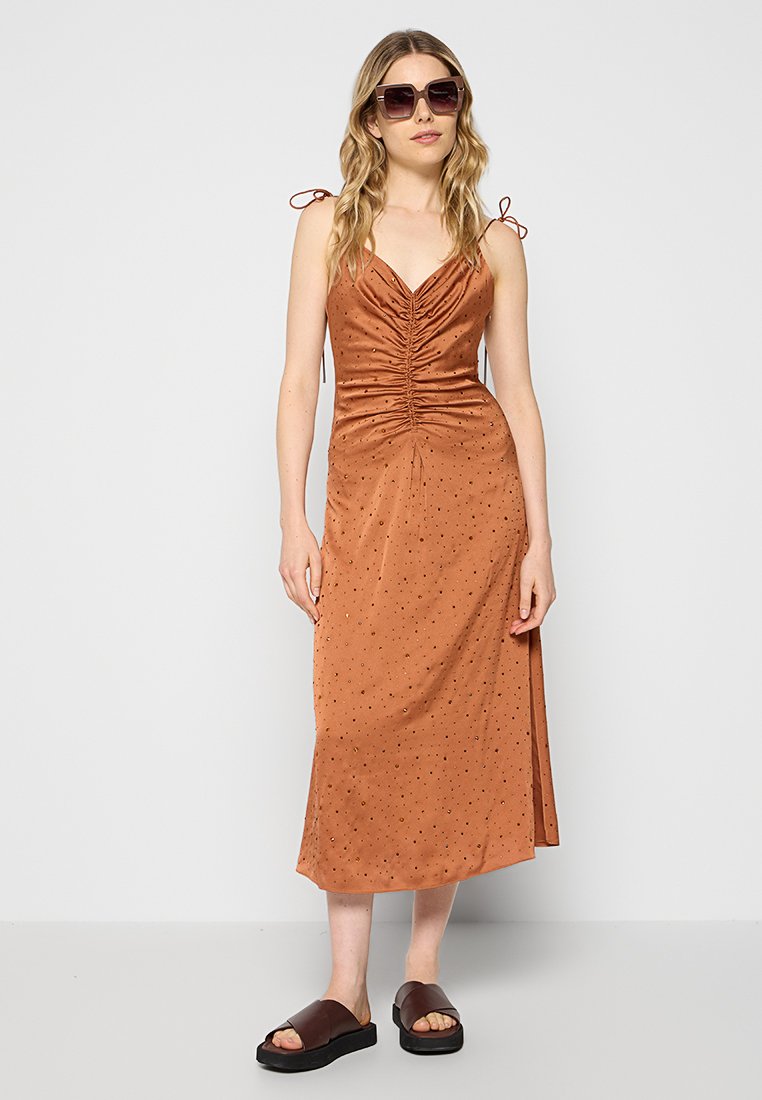 Vestido de satén marrón con fruncido en el busto, tirantes delgados ajustables y una abertura lateral. Presenta un patrón de puntos sutil. Combinado con sandalias oscuras.