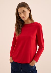 Röd långärmad topp i lättviktsmaterial, med rund halsringning och sidofickor. Enkelt designad med en slät yta.