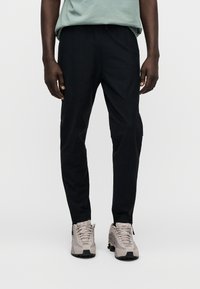 Calças pretas afuniladas com textura suave, apresentando punhos com fechos de correr. Combinadas com sapatos bege que têm uma superfície texturizada e solas escuras.