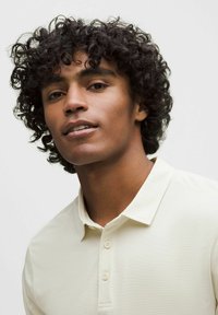 Polo shirt in lichtbeige met een kraag en drie knopen. De stof lijkt glad met een subtiele textuur. Het model heeft krullend haar.