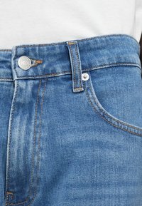 Εγγύς πλάνο της μέσης ενός μπλε τζιν παντελονιού με μεταλλικό κουμπί, θηλιά ζώνης, νίκελ και εμφανή πορτοκαλί ραφή πάνω σε φόντο λευκού πουκαμίσου.