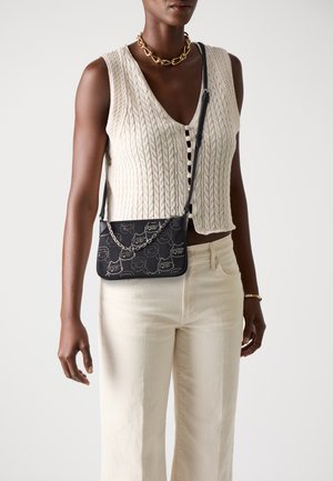 Black shoulder bag with a patterned design, featuring a silver chain strap. The model wears a cream knitted top and light beige pants.
