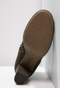 Botín de ante marrón con tacón de madera apilado, suela de goma texturizada y cremallera lateral, mostrado con la suela hacia arriba sobre un fondo blanco.