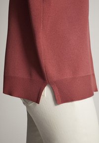 Rdzawy sweter z miękkiej dzianiny, z bocznymi rozcięciami u dołu i prążkowanymi detalami, w połączeniu z białymi spodniami.