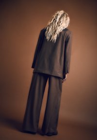 Hnedý oversized sako s tenkými prúžkami a široké nohavice s jemnou textúrou. Model stojí chrbtom, opretý o hnedé pozadie.