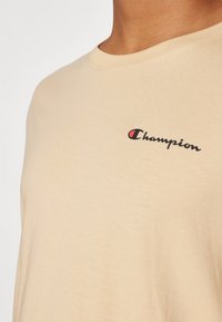 Beige színű pamut póló kerek nyakkivágással, fekete "Champion" logóval, piros kiemeléssel. Puha tapintású, laza szabású.