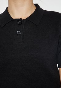 Černé pletené polo tričko s límečkem, které má tři knoflíky a krátké rukávy. Tkanina se zdá být měkká a strukturovaná.