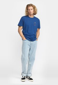 Camiseta de algodón azul con un pequeño gráfico colorido en el pecho. Combinada con jeans de mezclilla de lavado claro y zapatillas negras con acentos blancos.