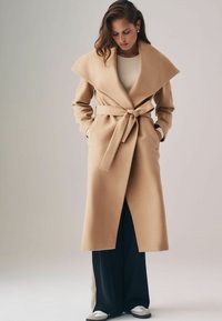 Beige gyapjú trench coat széles gallérral, övvel a deréknál és oldalsó zsebekkel, tengerészkék nadrág felett viselve.