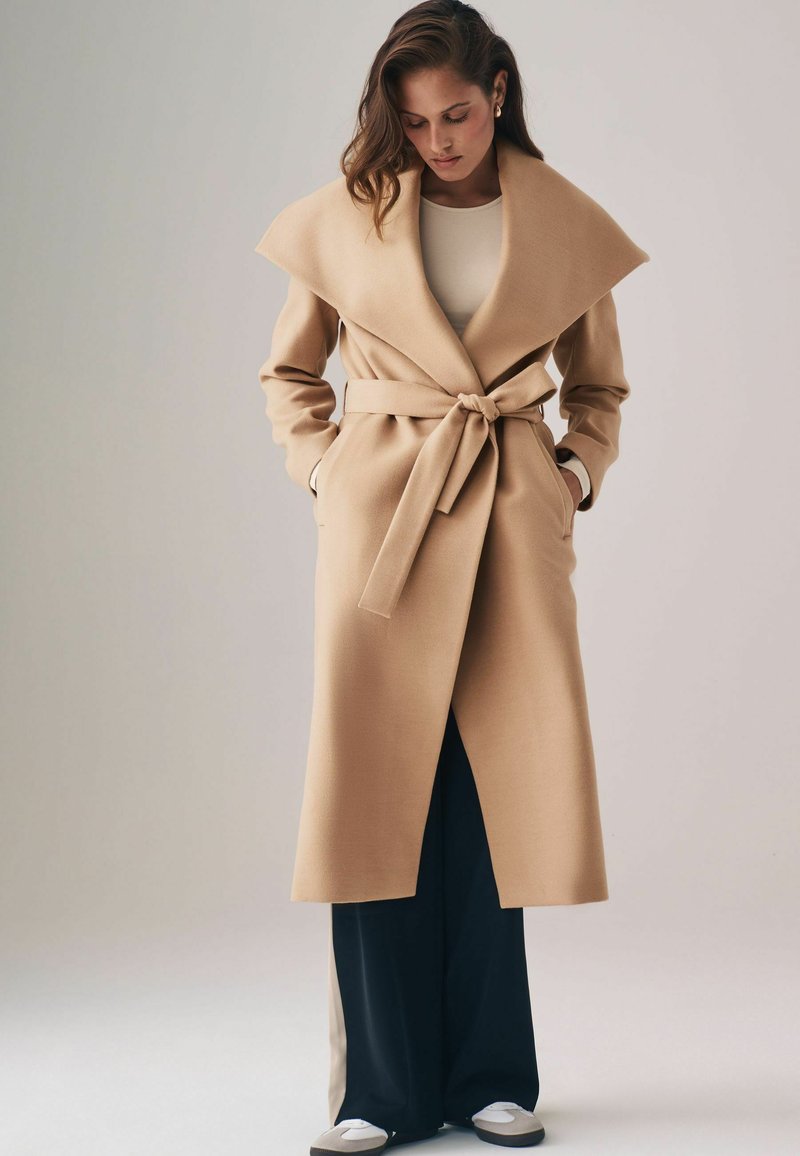 Beige Woll-Trenchcoat mit breitem Kragen, tailliertem Gürtel und Seitentaschen, getragen über dunkelblauen Hosen.