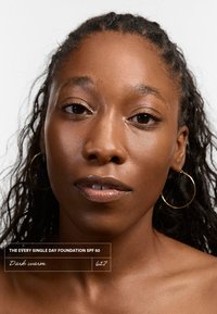 Every Single Day Foundation SPF 50, tono "Oscuro cálido" 627. El modelo tiene piel de tono marrón medio, cabello texturizado, pendientes de aro dorados y un brillo labial natural.