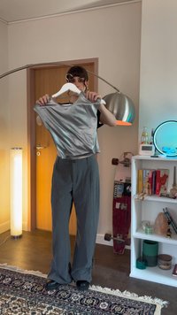 Person som står innendørs og holder en sølvfarget ermeløs topp på en kleshenger, med et skateboard og en bokhylle synlig i bakgrunnen.