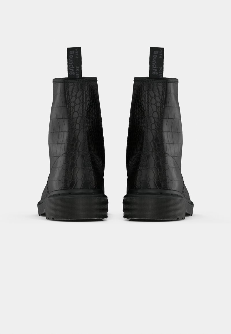 Un par de botas negras hasta el tobillo con un patrón texturizado de cocodrilo, tiradores negros que dicen "CON SUELAS REVOLUCIONARIAS" y suelas de goma gruesas, visto desde la parte trasera.