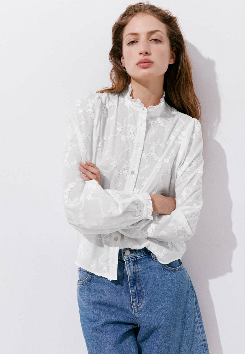Femme aux longs cheveux bruns portant un chemisier blanc en dentelle florale et un jean bleu, debout, les bras croisés sur un fond blanc.