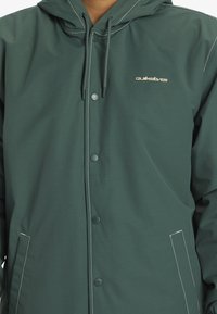 Veste vert foncé avec capuche, boutons-pression et accents de couture blanche. Fabriquée en matériau lisse et résistant à l'eau, avec un logo sur la poitrine.