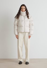 Puhast bela jakna z visokim ovratnikom, z vodoravnimi kviltami. V kombinaciji z ohlapnimi smetanastimi hlačami in temnim obutvijo.