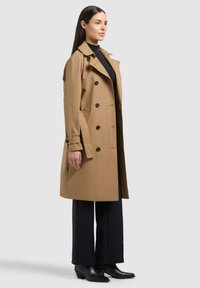 Donna con lunghi capelli scuri che indossa un trench color camoscio doppiopetto, dolcevita nero, pantaloni a gamba larga e stivaletti neri, vista di lato.