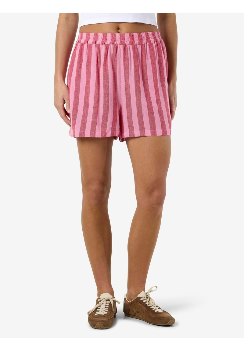 Kvinde iført pink og røde shorts med lodrette striber, elastisk talje og brune snørelåssko, stående med afslappede arme.