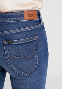 Närbild av blå denimjeans med bakficka och ett brunt lädermärke märkt "Lee" på midjebandet.