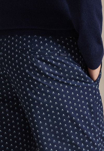 Námořnické kalhoty s malým bílým vzorem lebky a zkřížených kostí, zadní klopa kapsy s nápisem "RLX", nošené s tmavým svetrem a rukou v boční kapse.