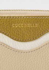 Detailní záběr na texturovanou koženou taštičku s prohnutými švy v béžové, bílé a olivové barvě, s vyraženým zlatým logem "COCCINELLE" a zlatým zipem.