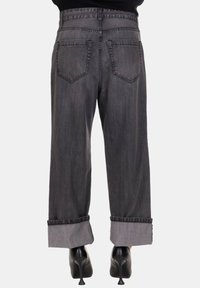 Persona che indossa jeans a gamba larga grigio scuro con risvoltini e scarpe con tacco nere, in piedi contro uno sfondo neutro.