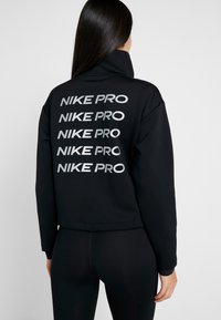 Zwarte Nike Pro cropped sweatshirt met een hoge kraag en meerdere gestapelde zilveren logo's op de achterkant. Glad textuur en een ontspannen pasvorm.