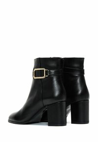 Bottines en cuir noir avec un bout carré, un talon épais et haut, et une boucle en or décorative sur la sangle de la cheville. Texture lisse.