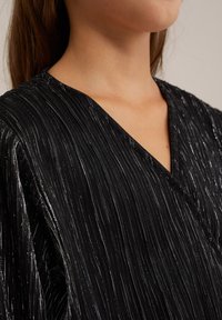 Svart V-ringad topp med en texturerad, ribbad tyg som har metalltrådar, vilket skapar en glänsande effekt över ytan.