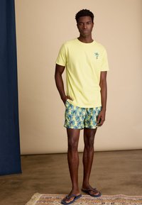 Heldergele katoenen t-shirt met een klein palmbomenteken; blauwgele gebloemde zwembroek met palmbomen; donkerblauwe slippers.