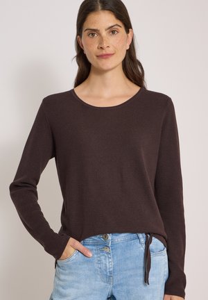 Femme aux longs cheveux foncés portant un haut marron à manches longues et un jean bleu clair, debout avec une main dans la poche sur un fond uni.