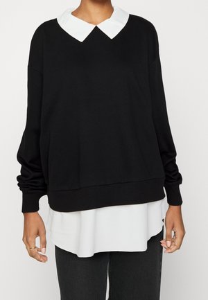 Persona vistiendo una sudadera negra con el cuello y el dobladillo de una camisa blanca asomando por debajo, combinada con pantalones negros.