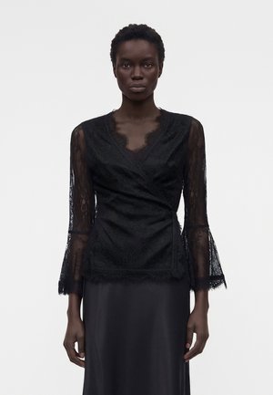 Γυναίκα που φοράει μαύρο δαντελένιο τοπ τύπου wrap με διαφανή μακριά μανίκια και κυματιστό ντεκολτέ, συνδυασμένο με μαύρη σατέν φούστα, στέκεται μπροστά σε απλό φόντο.