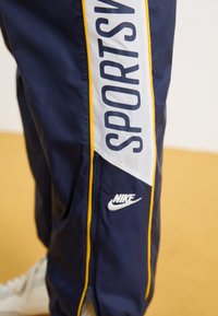 Pantaloni sportivi blu navy con pannelli laterali bianchi e gialli, caratterizzati da testo "SPORTS" e logo Nike. Inclusa la cerniera all'orlo.