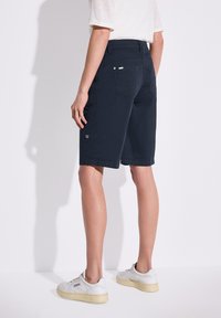 Pantaloni cargo blu navy con tasche laterali, realizzati in tessuto leggero; presentano una vestibilità regolare e orli arrotolati. Indossati con sneaker bianche.