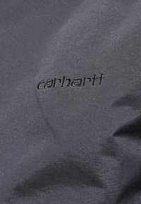 Un tessuto nero con una superficie liscia presenta un logo "carhartt" in rilievo, posizionato diagonalmente vicino al centro dell'immagine.