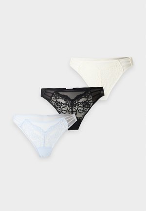 Femme debout pieds nus portant un soutien-gorge en dentelle noire et une culotte assortie sur un fond clair uni.