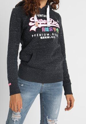 Person trägt einen dunkel gesprenkelten Hoodie mit buntem "Superdry"-Schriftzug und abgenutzte hellblaue Jeans, steht vor einem einfarbigen Hintergrund.