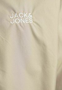 Ljusbeige tyg med en subtil rutad textur. Har en broderad "JACK & JONES"-logotyp i vitt. Enkel design med minimal detaljering.