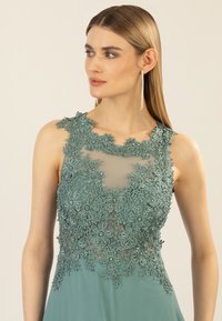 Sleeveless dress in muted teal, featuring intricate floral lace appliqué at the neckline and beaded accents on a sheer overlay.