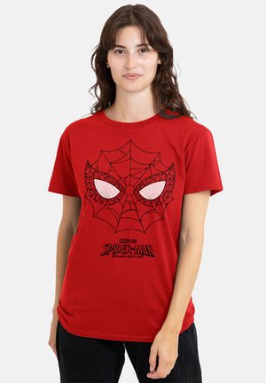 Young woman wearing a red Spider-Man T-shirt with a web and eye design and black pants, standing against a plain light background.
